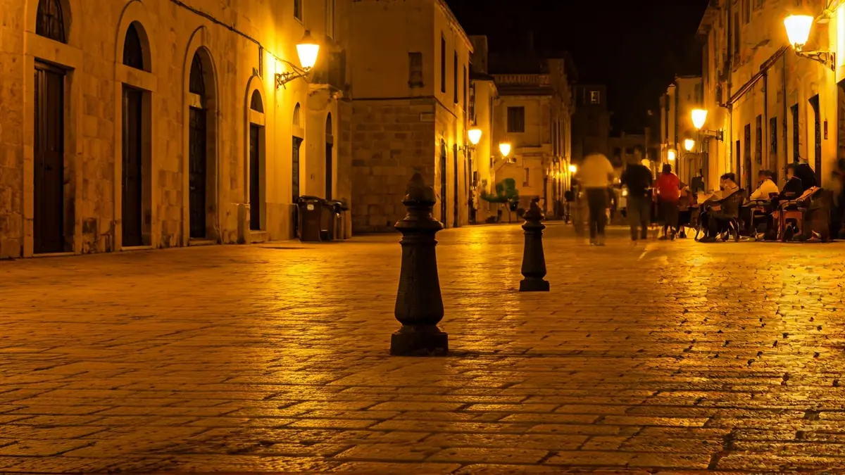 Imatge genèrica d'una plaça mediterrània de nit, amb fanals i figures borroses al fons.