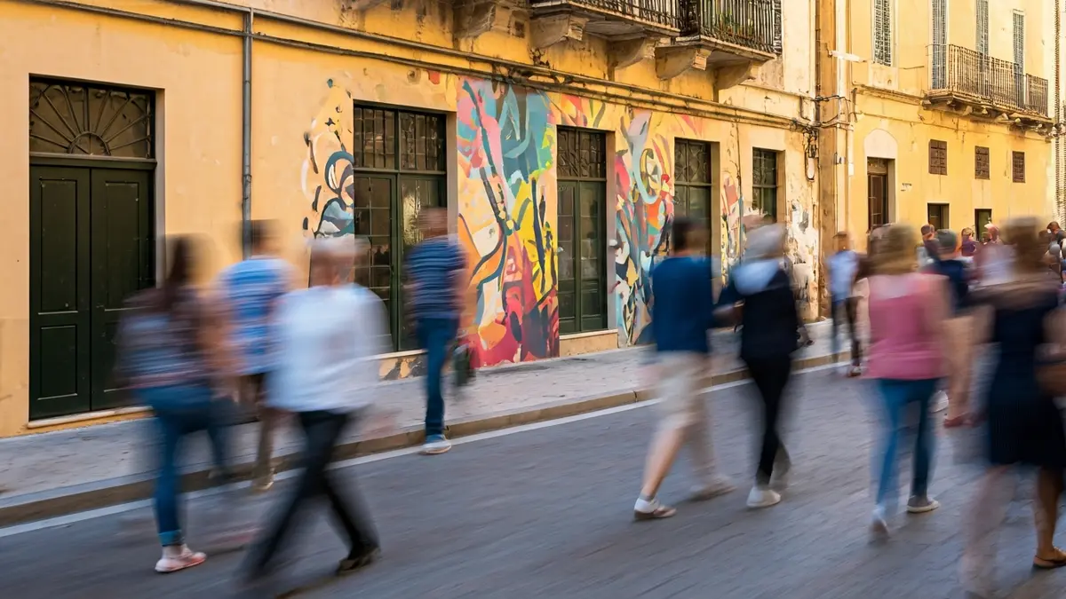Image of an artistic mural on a building facade in a Mediterranean city, with blurred figures of people and a festive atmosphere.