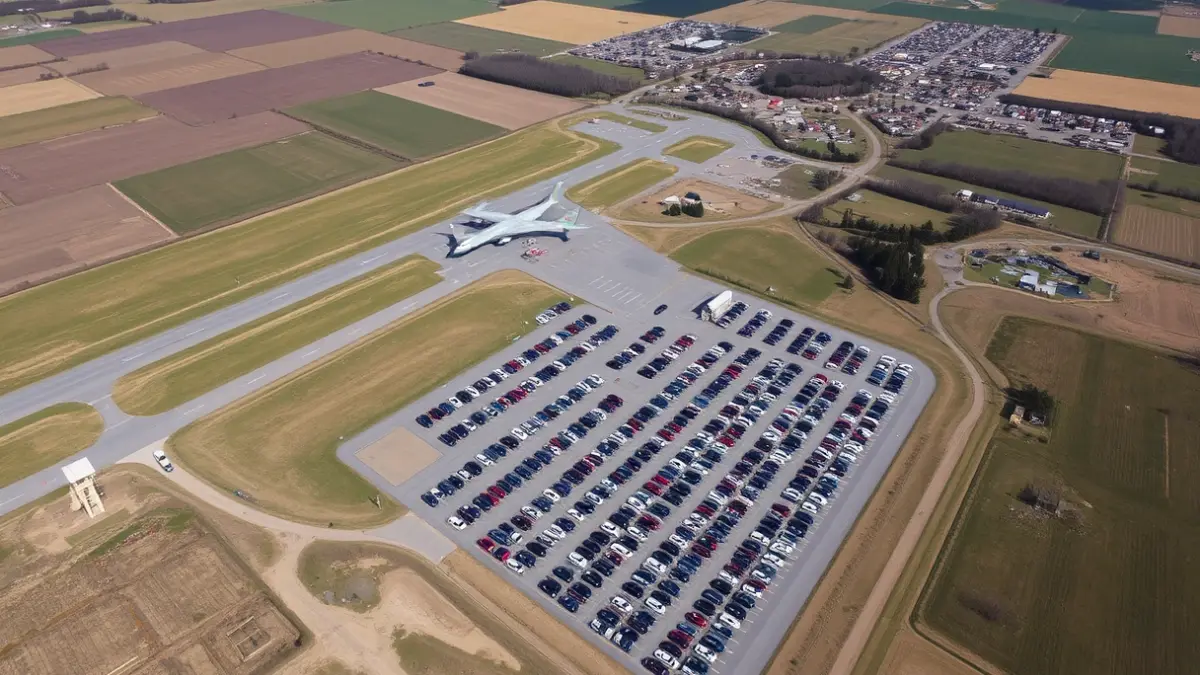 Aerial image of a large parking lot full of vehicles near an airport, surrounded by rural landscape and some scattered houses.