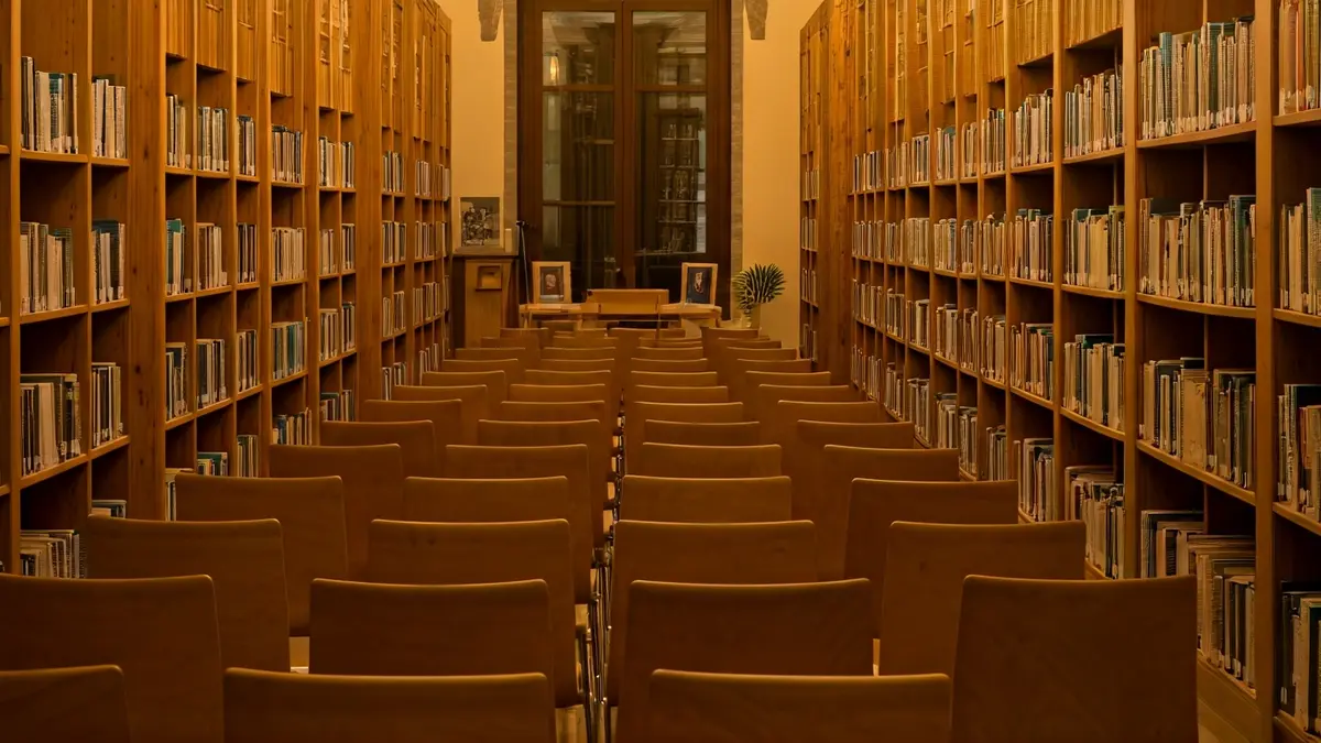 Imagen de una sala de exposiciones o biblioteca con luz cálida, estanterías de madera y un podio con micrófono, en un edificio de estilo mediterráneo.