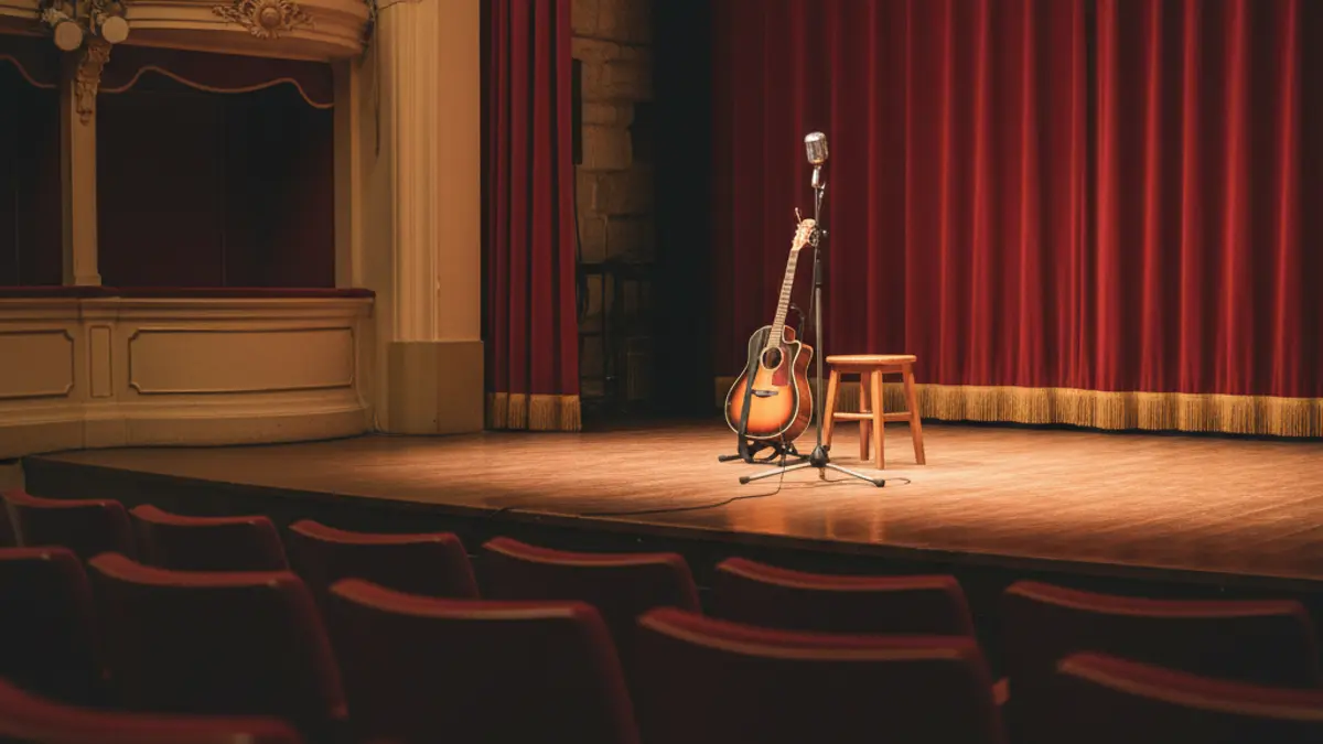 Generic image of a theater stage with a microphone and a guitar, warm lighting.